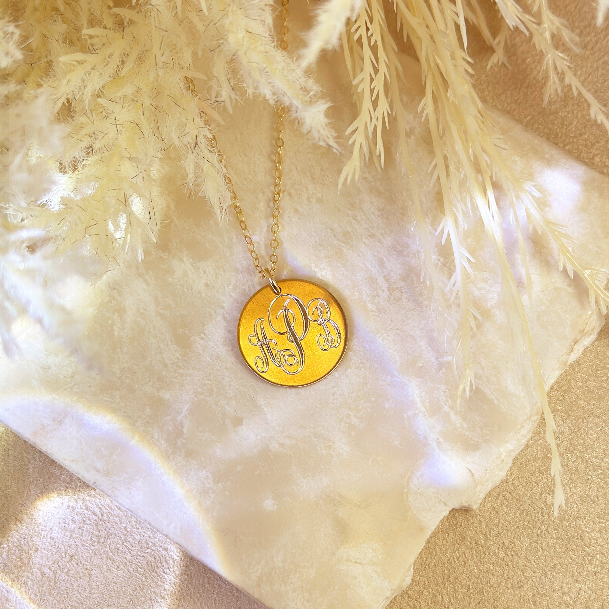 Gold monogram disk necklace with engraved script initials on a delicate chain, displayed on soft neutral fabric and pampas grass – personalized jewelry gift for her, girlfriend, student, or new job celebration, milestone gift