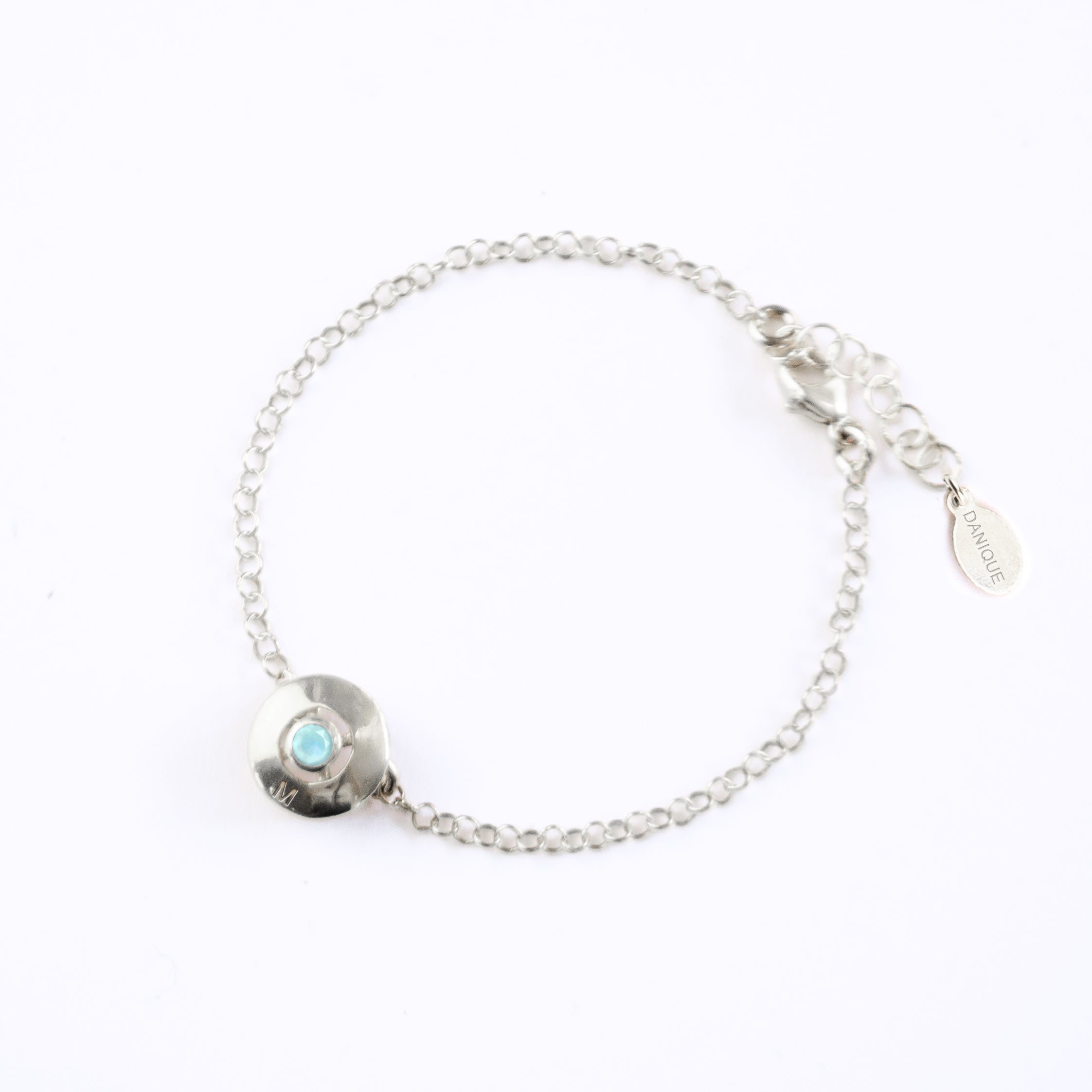 A 925 sterling silver donut bracelet with an aqua chalcedony stone and the letter "M" engraved on the bottom of the disc rests against a white background. This bracelet is hypoallergenic and is sure to make the day of anyone who receives this as a gift! This is the perfect thing for Mother's Day!
