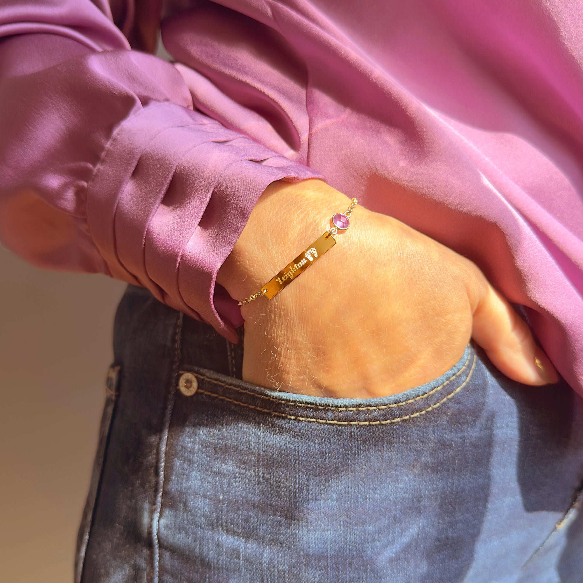 Dainty personalized baby footprint birthstone bracelet in yellow gold filled, styled on wrist. Features custom engraving and birthstone charm. Ideal push present, Mother’s Day jewelry, or new mom gift. Handcrafted adjustable bracelet in sterling silver, gold, or rose gold.