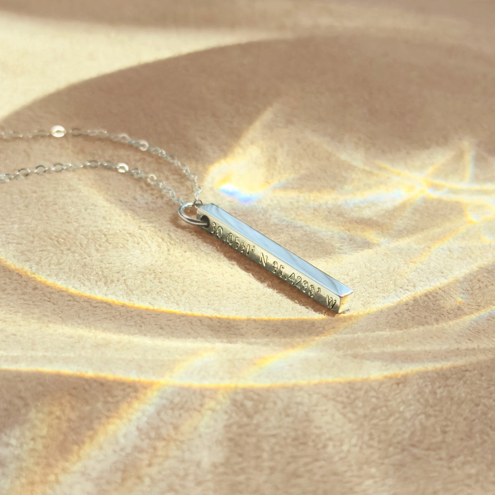 A minimalist sterling silver bar pendant etched with exact latitude longitude necklace details, styled on a soft backdrop with prism light. This slim GPS coordinates necklace turns a special place—first date, proposal spot, new home—into elegant location jewelry. Deep laser text runs along one face of the bar for a modern engraved coordinate bar look; perfect as a travel keepsake gift or anniversary memento with custom map coordinates and adjustable chain length.