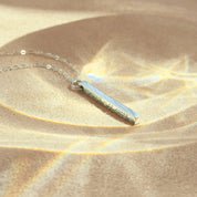 A minimalist sterling silver bar pendant etched with exact latitude longitude necklace details, styled on a soft backdrop with prism light. This slim GPS coordinates necklace turns a special place—first date, proposal spot, new home—into elegant location jewelry. Deep laser text runs along one face of the bar for a modern engraved coordinate bar look; perfect as a travel keepsake gift or anniversary memento with custom map coordinates and adjustable chain length.