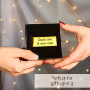 Hands presenting a gift box that holds a gold engraved money clip with the cheeky message “Cash me if you can.” The sleek metal clip sits on a black velvet insert against warm fairy-light bokeh, emphasizing ready-to-gift packaging. Personalized money clip for men, ideal birthday, Christmas, graduation, or Father’s Day gift for dad, husband, or boyfriend. Slim wallet alternative with custom text or handwriting engraving, handcrafted by Danique Jewelry.