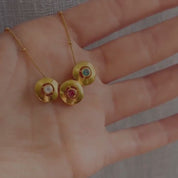A hand holds a 3-charm donut birthstone necklace on a gold satellite beaded chain. In the second frame, a person shows off a 1-charm donut bracelet with a kyanite september birthstone in the center. In the third frame, a person in a black shirt wears a gold 2-charm donut birthstone necklace with a moonstone and a ruby. The final two frames show a 1-charm moonstone necklace and a 1-charm ruby bracelet on a person, each set in gold-fill. Personalize these necklaces and bracelets to fit your family!