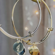 A silver and a gold birthstone and disc bangle bracelet. Personalized name and gemstone bangle, custom mother's jewelry, half bezel teardrop prong bracelet. A person wears a gold bangle with citrine and engraved name Oliver in Classic font. Two hands move a rose gold charm bracelet back and forth. A person wears a gold charm bracelet in the sun. A silver bangle in an open red Danique Jewelry box.