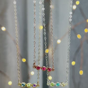 3 custom bar necklaces pan down. A person wears a triple ruby bar necklace. Two hands hold a three birthstone pendant up close to show the bezel setting and the quality facets. Two hands hold an open red Danique Jewelry box with a silver triple gemstone horizontal bar necklace.