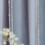 The camera pans down to reveal two round cascade necklaces in silver and gold. A person in a white shirt with long brown hairs wears a gold round cascade necklace with citrine quartz and ruby quartz suspended on a gold filled cable chain. The person thumbs a 2-stone necklace, showing off the faceted sparkling birthstones. A person shows off a three stone silver cascade necklace with blue and green stones in an open red danique jewelry box.