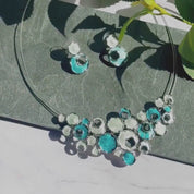 A blue and green Boho connected circles bubble necklace and earring set sits on a granite background. A panover of simple circle station necklaces show color combinations of yellow cream and green and orange blue and teal. A panover of matching earrings follows. An abstract flower necklace and matching earrings appear next. This Boho collection is perfect for those who want to add a unique mix of shapes and colors to their repertoire, and can be crafted in custom colors for an extra personalized feel.