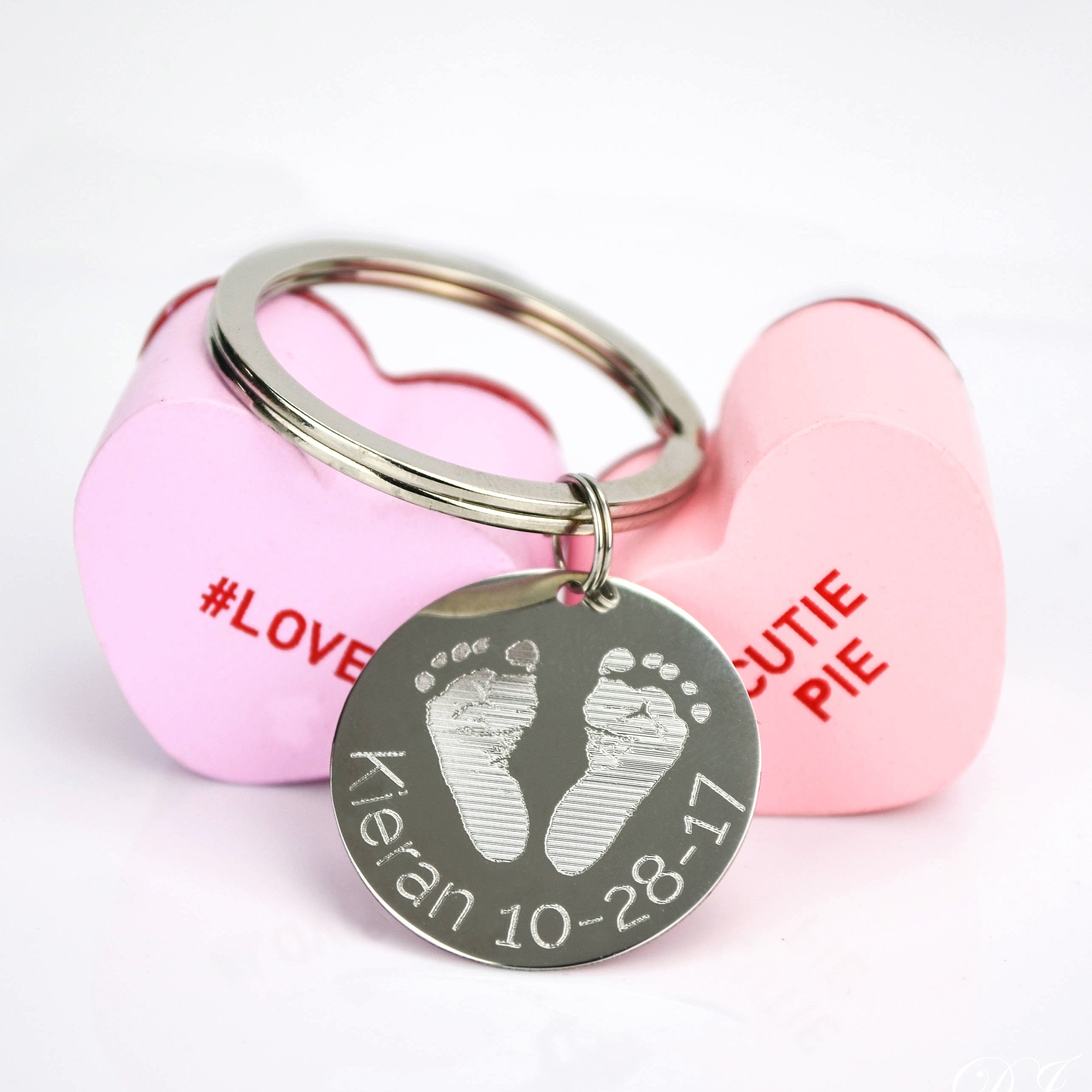 Single footprint keychain engraved in block font with baby's name and birth date. Sitting on valentine's hearts.