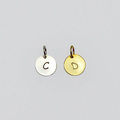 Polished disk hand stamped initial charm, initial oval gold silver, rose, on white background.