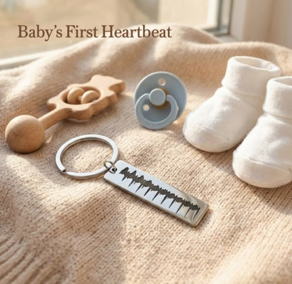 Silver baby heartbeat sonogram keychain on a soft knit blanket beside white baby booties, a pacifier, and wooden rattle, styled in warm sunlight with text reading Baby’s First Heartbeat.