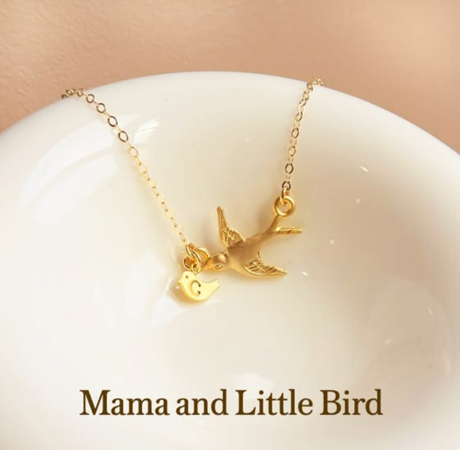 Gold mama and baby bird necklace with a tiny initial charm displayed on a white dish, styled in soft warm light with elegant text, symbolizing motherhood, love, family, and connection.