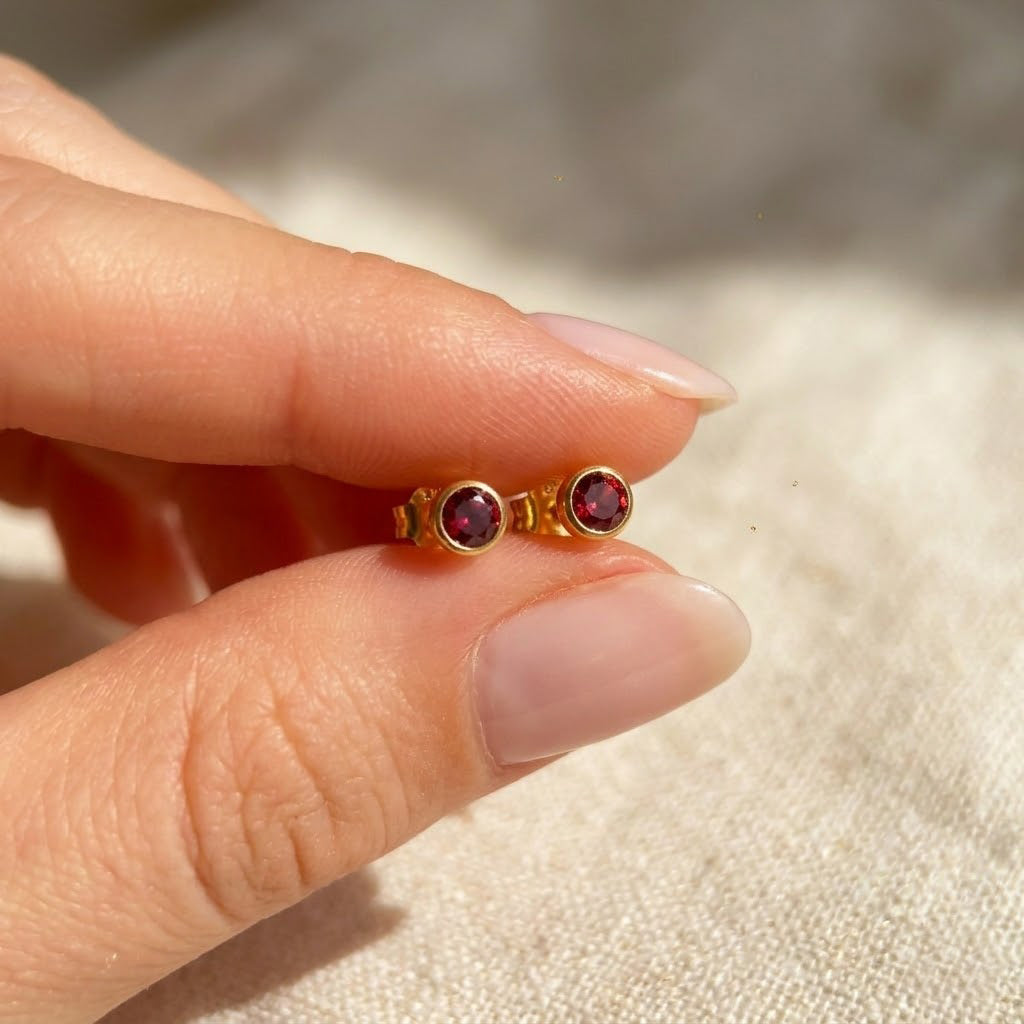 Hand holding tiny birthstone stud earrings showing small scale and delicate bezel set gemstone design