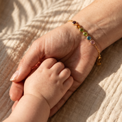 A delicate gold bracelet with colorful gemstone charms worn on an adult wrist, holding a baby's hand in warm sunlight.