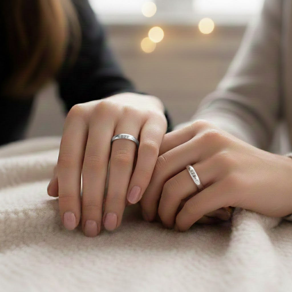 Two people resting hands together on a soft blanket, each wearing a slim silver engraved ring, cozy lifestyle photo showcasing matching personalized bands, custom name or message rings for best friends, sisters or couples, meaningful promise ring gift for her, minimalist everyday jewelry.