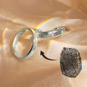 Sterling silver fingerprint ring set with deep wrap-around engraving of a real fingerprint on a polished band, shown on a blush satin background with soft rainbow light reflections; custom engraved wedding band or memorial ring that can be personalized with your loved one’s fingerprint and inside message, meaningful keepsake gift for him or her.