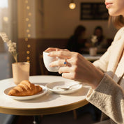 Dainty gold stackable birthstone ring with vibrant blue gemstone worn on a woman’s hand as she holds a coffee cup in a cozy café, soft morning light and bokeh lights in background, croissant and minimal table setting, thoughtful everyday mothers ring gift.
