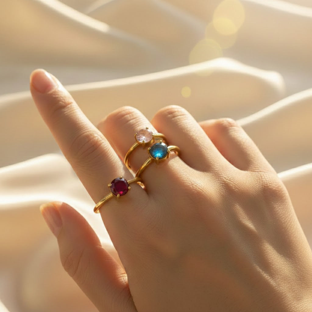 Hand wearing three stackable birthstone rings in 18K gold vermeil, each dainty bezel-set gemstone in rich jewel tones, styled as a minimalist mothers ring set on a soft champagne satin background; customizable birthstones for kids or grandkids, perfect sentimental jewelry gift for mom or grandma, birthday, anniversary or Mother’s Day, showing how these slim stacking bands mix with engagement, wedding or promise rings and layered everyday gold jewelry looks.
