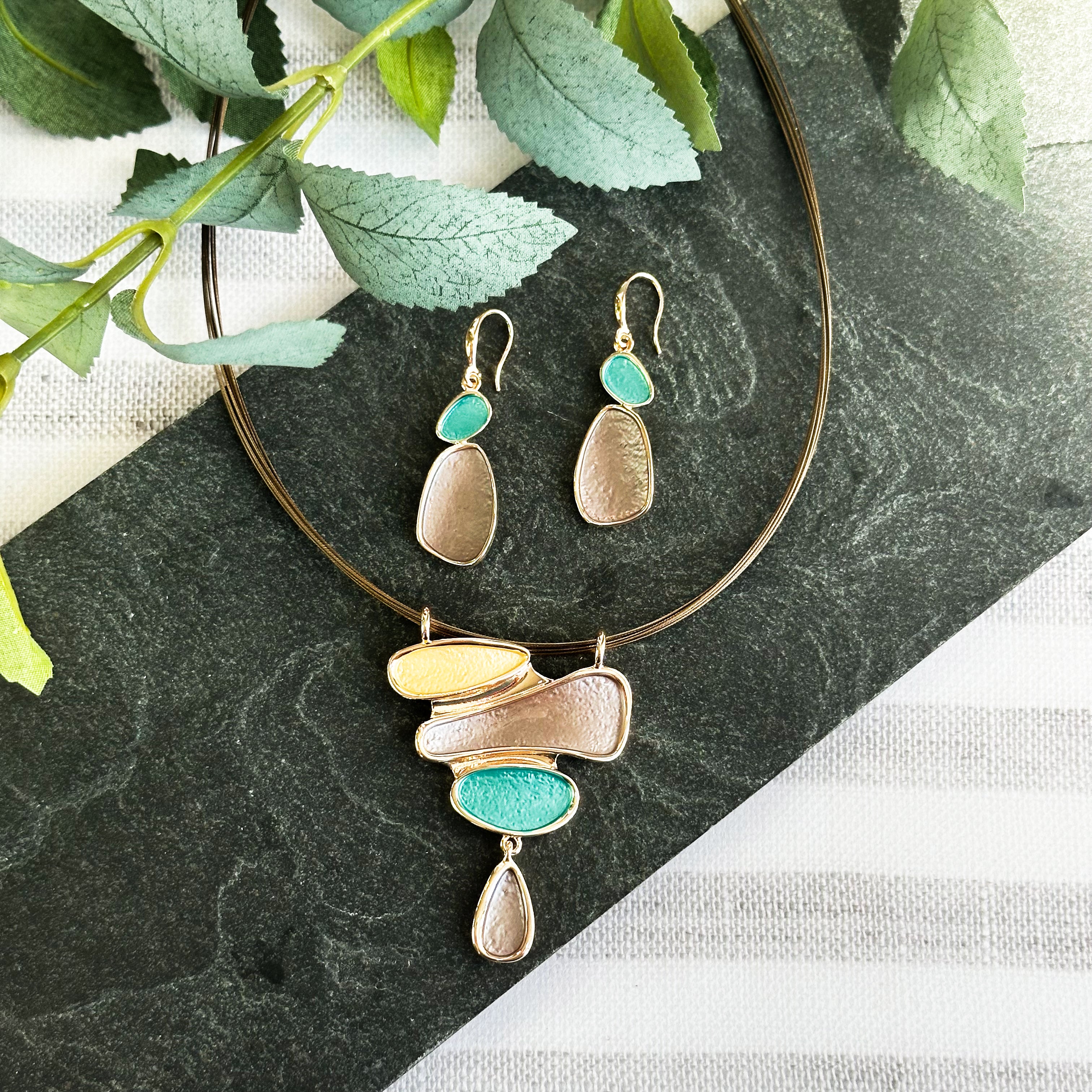 Matching Earring and Necklace Set. Earrings are dangle earrings with two oval shapes painted in teal and brown. Necklace is a pendant with three organic shapes and a dangling teardrop painted in gold, brown, and teal. Necklace has a copper wire chain