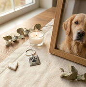 Custom pet paw print and name keychain displayed beside a framed photo of a golden retriever, styled as a heartfelt memorial keepsake. Personalized stainless steel dog paw keychain engraved with pet name “Bella,” shown on a neutral linen surface with candle and greenery accents. Meaningful pet remembrance gift for dog lovers, pet parents, or those honoring a beloved dog. Thoughtful sympathy gift, pet loss memorial, or everyday key accessory celebrating unconditional love.