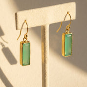 Aqua chalcedony baguette bar earrings in gold displayed on a cream jewelry stand in natural sunlight.