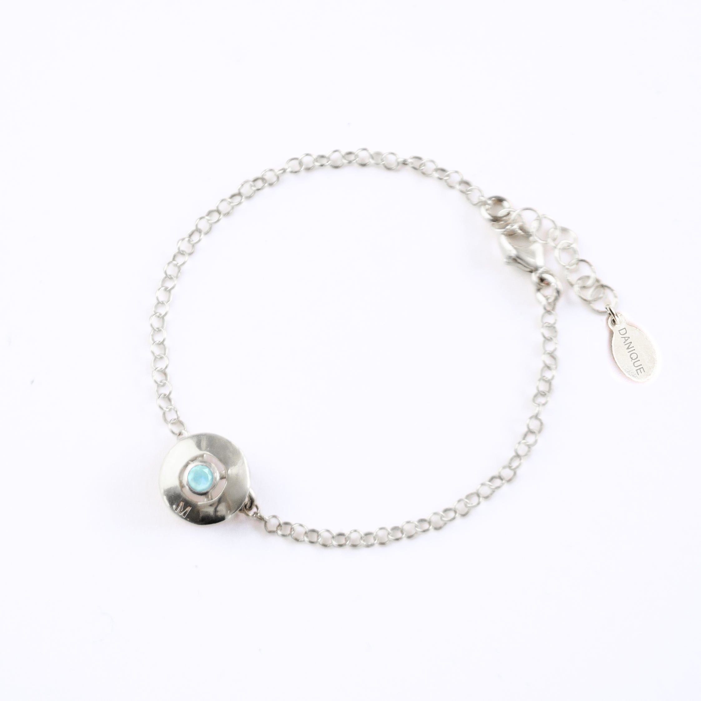 A 925 sterling silver donut bracelet with an aqua chalcedony stone and the letter "M" engraved on the bottom of the disc rests against a white background. This bracelet is hypoallergenic and is sure to make the day of anyone who receives this as a gift! This is the perfect thing for Mother's Day! 