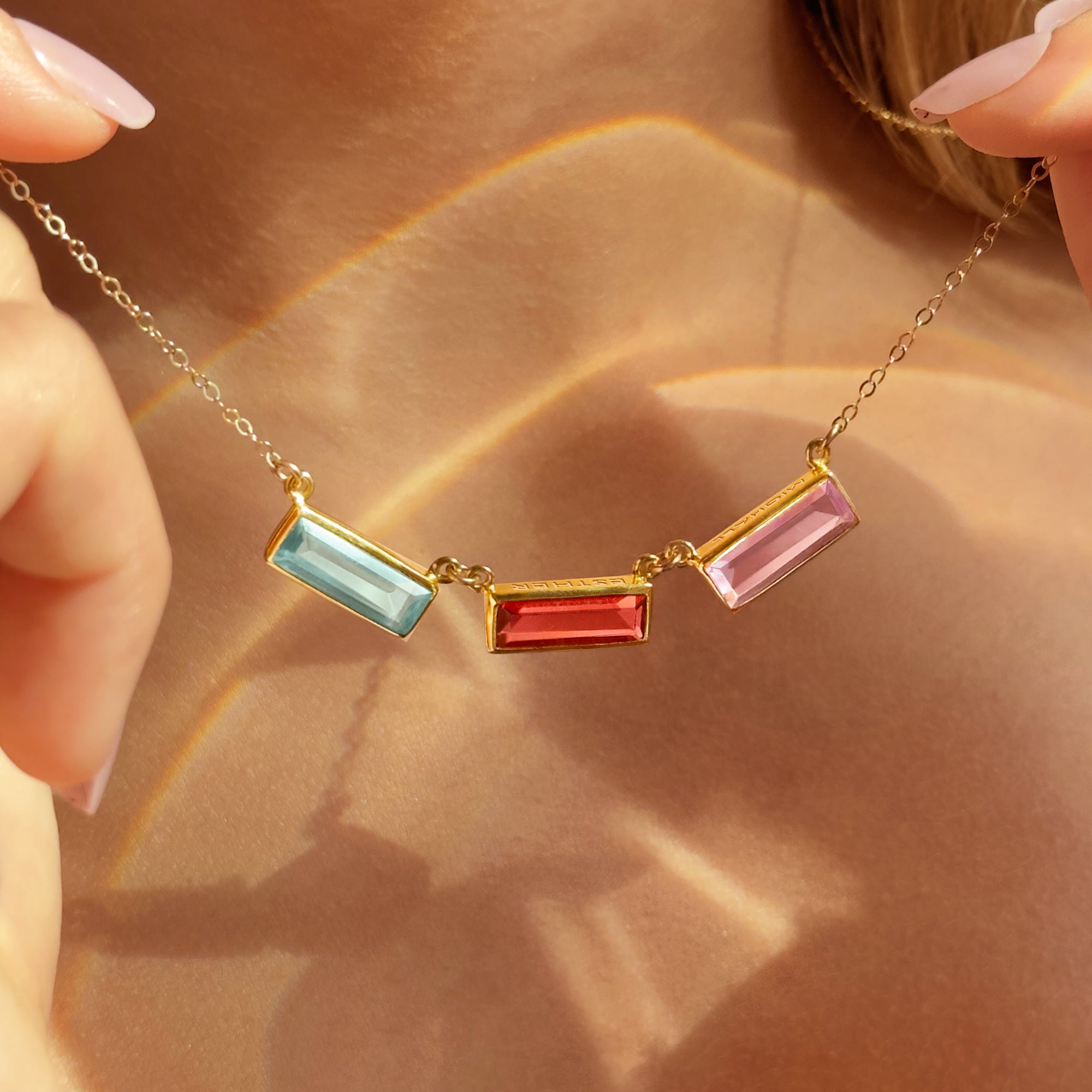 Adira baguette birthstone bar necklace for mom featuring three rectangular bezel-set gemstones on a delicate gold chain. Personalized engraving available with names, initials, dates, or a hidden message. Shown layered for everyday wear, this custom birthstone mom necklace is a meaningful gift for Mother’s Day, birthdays, or Christmas. Stones pictured include aquamarine, ruby, and rose hues; also available in sterling silver and rose gold options for grandma or new moms.