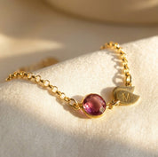 Detailed product photo of gold birthstone bracelet with engraved leaf initial charm, pink faceted stone, extender chain, and minimalist custom bracelet design