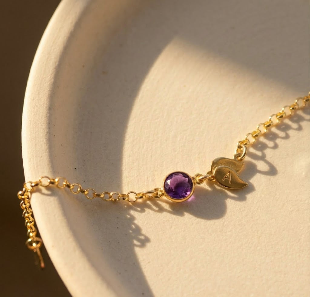 Close-up of personalized birthstone bracelet with pink gemstone and engraved initial leaf charm, adjustable gold bracelet with dainty chain and lobster clasp