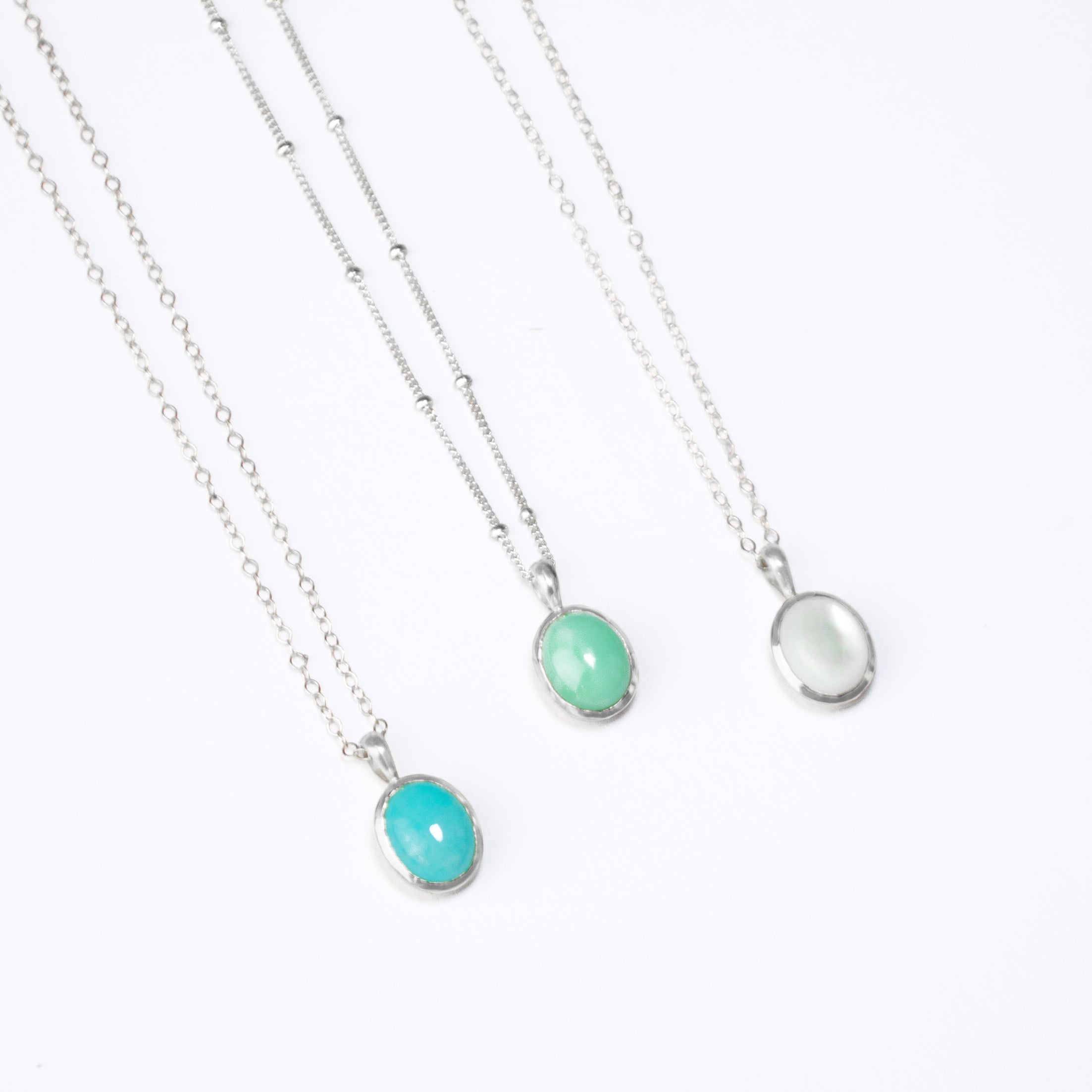 Three 925 Sterling Silver Oval Gemstone Pendant Necklaces on a white background. Silver Blue Amazonite Oval Pendant on a cable chain, Silver Mint Chrysoprase Oval Pendant on a Satellite Chain, and Silver Mother of Pearl Oval Pendant on a Cable Chain