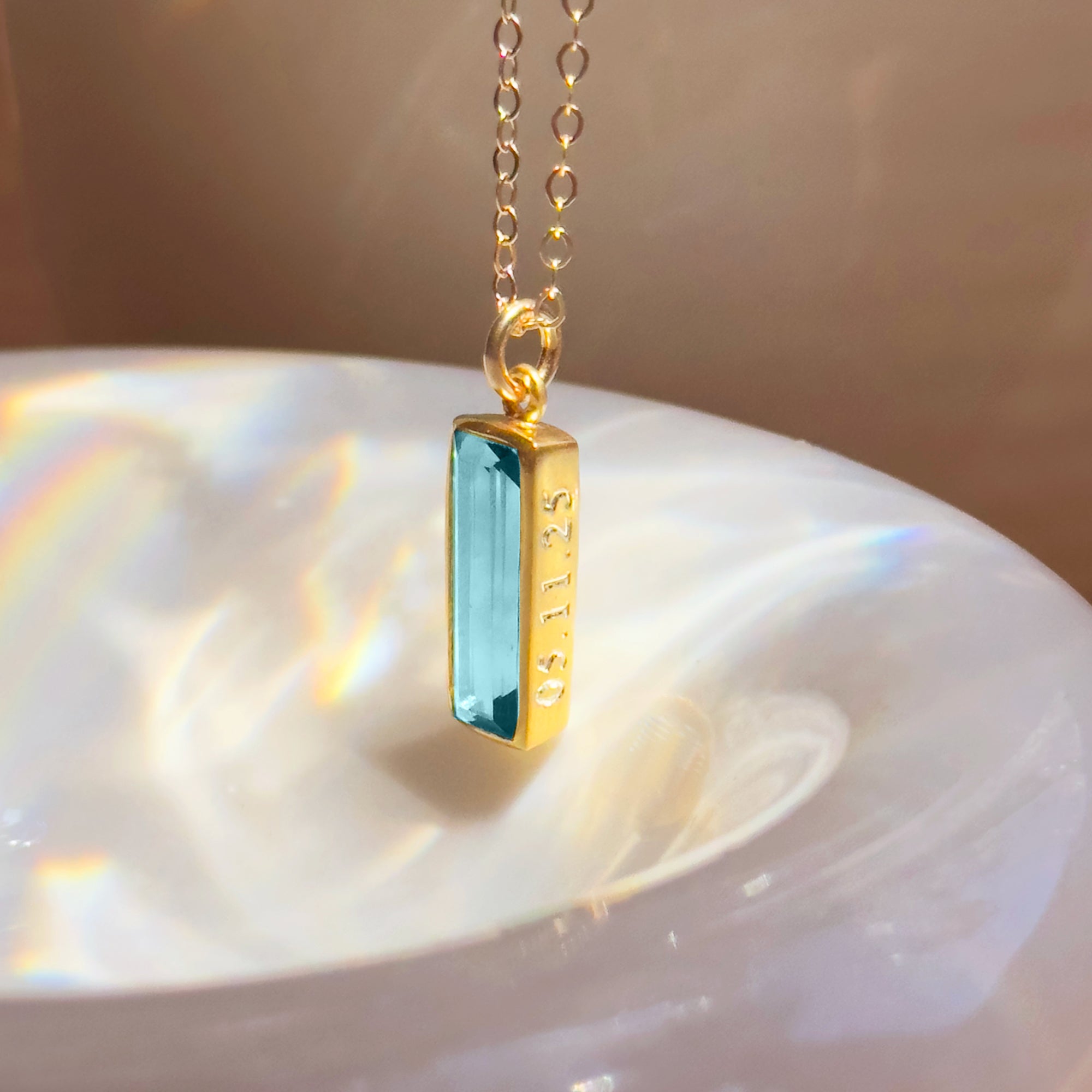 blue topaz bar necklace in gold vermeil shown as a close-up on soft ivory satin fabric, featuring a faceted rectangular december birthstone set in a vertical bezel pendant on a delicate chain, showcasing bright sky-blue sparkle and minimalist design, customizable as an engraved birthstone necklace for birthdays or holiday gifting, handmade sterling silver or gold vermeil gemstone jewelry ideal for mom, daughter, wife, or best friend