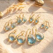 Faceted blue topaz earrings in seven teardrop, round, and geometric styles on cream fabric.