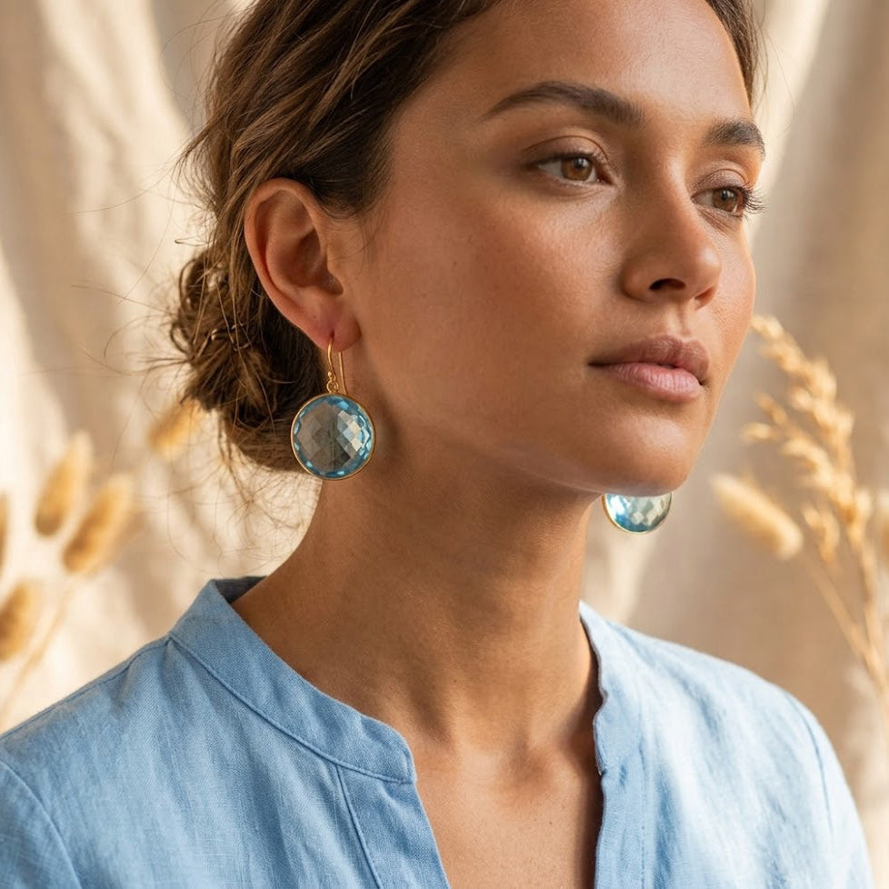 Woman wearing large round faceted blue topaz drop earrings in a yellow gold finish.