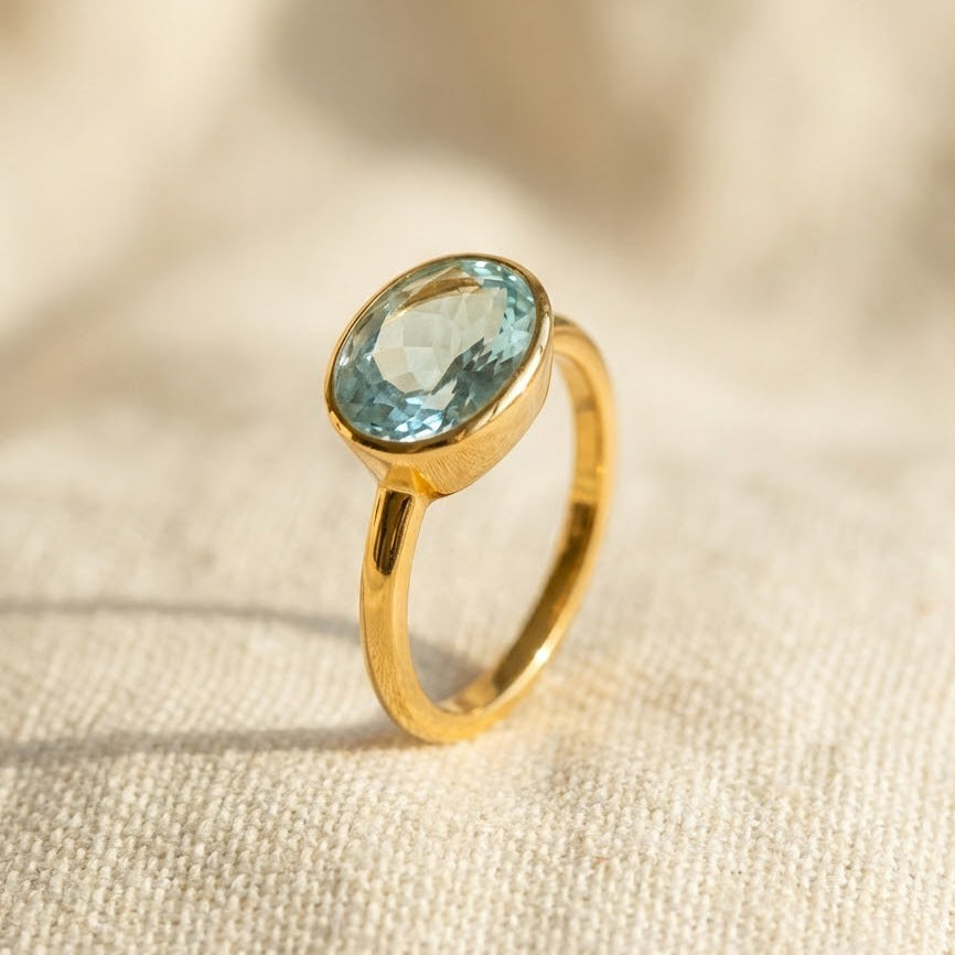 Gold oval blue topaz ring standing upright on neutral background, faceted December birthstone gemstone in bezel setting, elegant minimalist jewelry