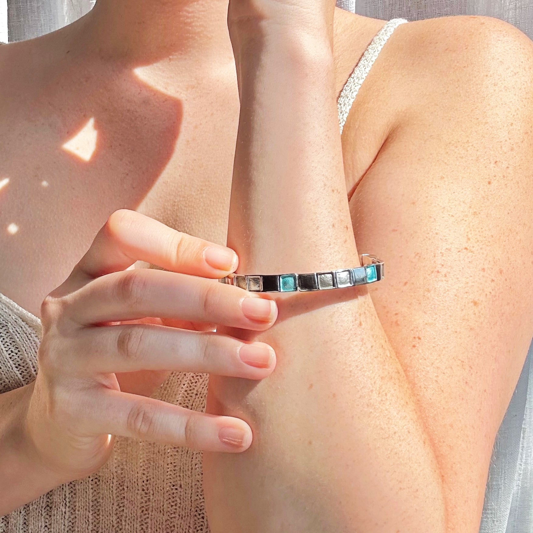 Rhodium plated cuff bracelet with a magnetic closure with square indents painted in silver, gray, and teal enamel is worn by a model in a white knit tank top