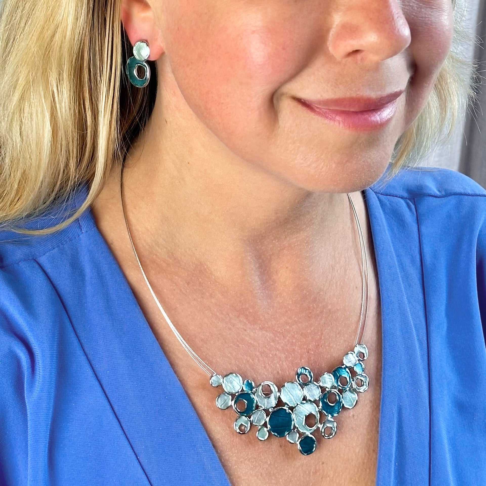 Matching Earrings and Necklace worn by a model with a steel cable chain with an organic triangular shaped pendant made up of rhodium plated circles painted in blue and silver enamel. Some of the circles contain a circular cut out.