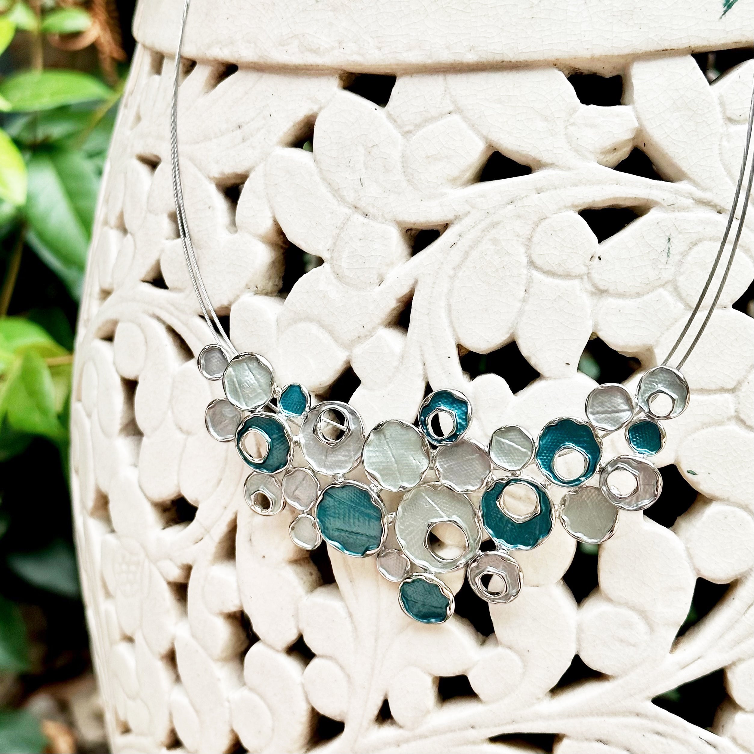 Necklace against a white ceramic pot made of interlocking plant shapes with a steel cable chain with an organic triangular shaped pendant made up of rhodium plated circles painted in blue and silver enamel.