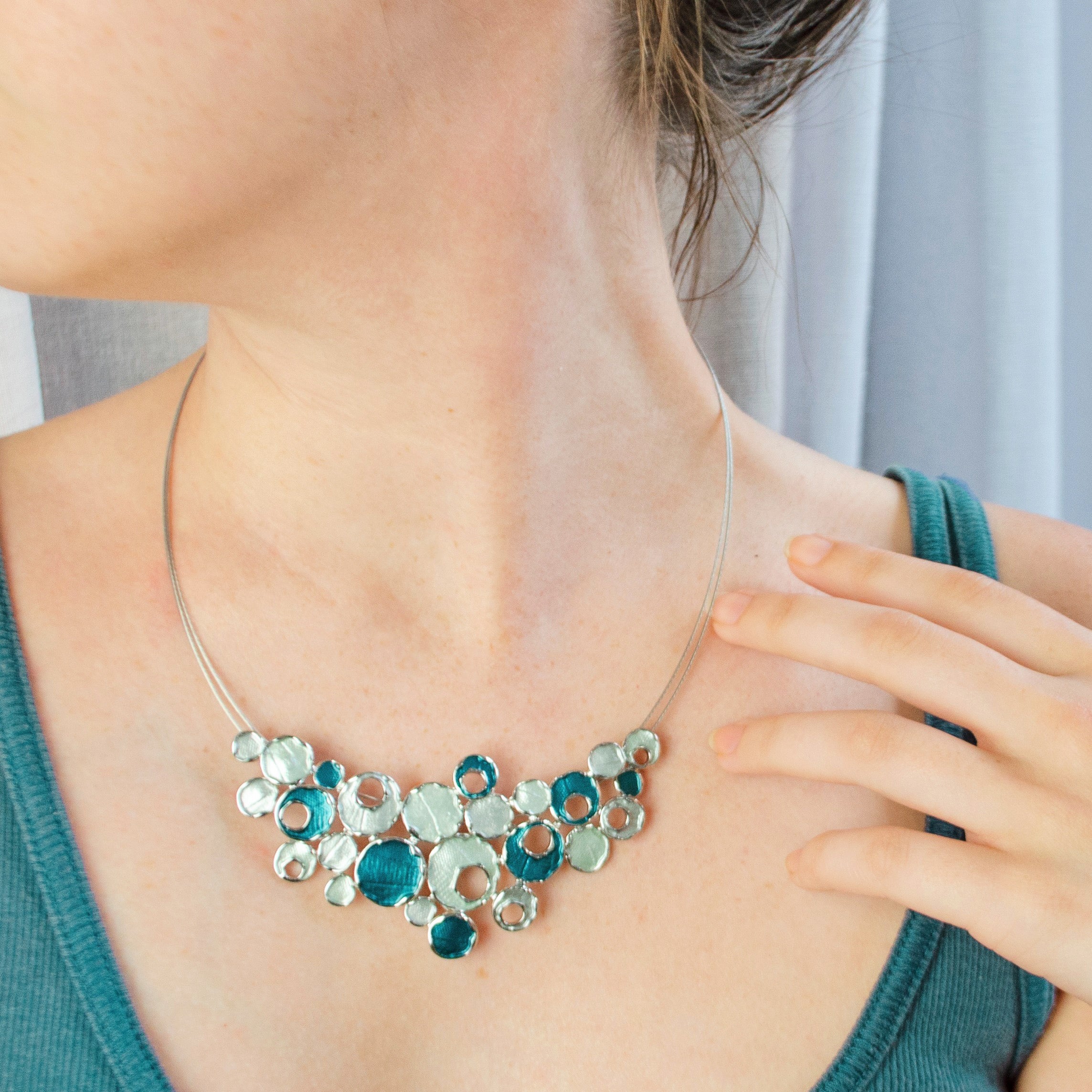 Necklace worn by a model with a steel cable chain with an organic triangular shaped pendant made up of rhodium plated circles painted in blue and silver enamel. Some of the circles contain a circular cut out.