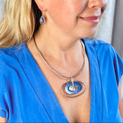 A pair of rhodium plated open oval drop earrings with hook backings and a textured oval disc at the center painted in green enamel are worn by a model with her head turned to the side and her hand by her jawline