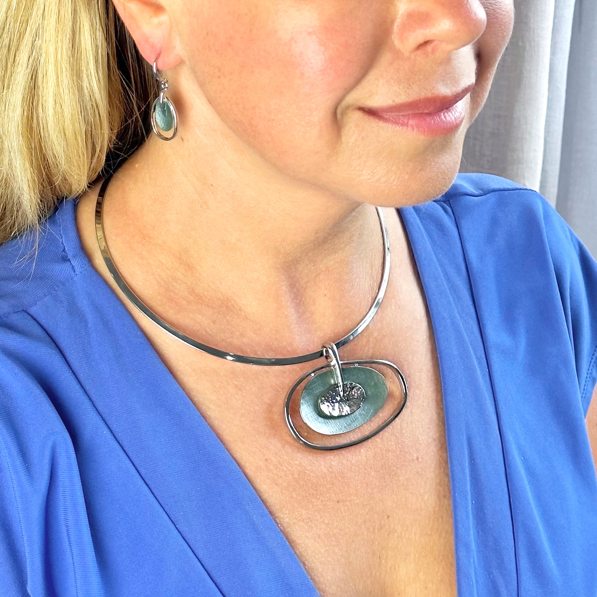 Rhodium plated Open Oval Pendant with two textured overlapping oval disks in the center, painted in green enamel and sitting on round choker with matching hook earrings worn by model in a blue blouse