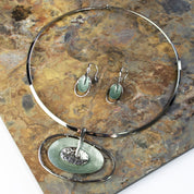 rhodium plated round choker with an Open Oval Pendant with two textured overlapping oval disks in the center surrounded by an oval cutout, the bottom disk is painted in green enamel. Necklace rests with matching earrings on a black stone tile.