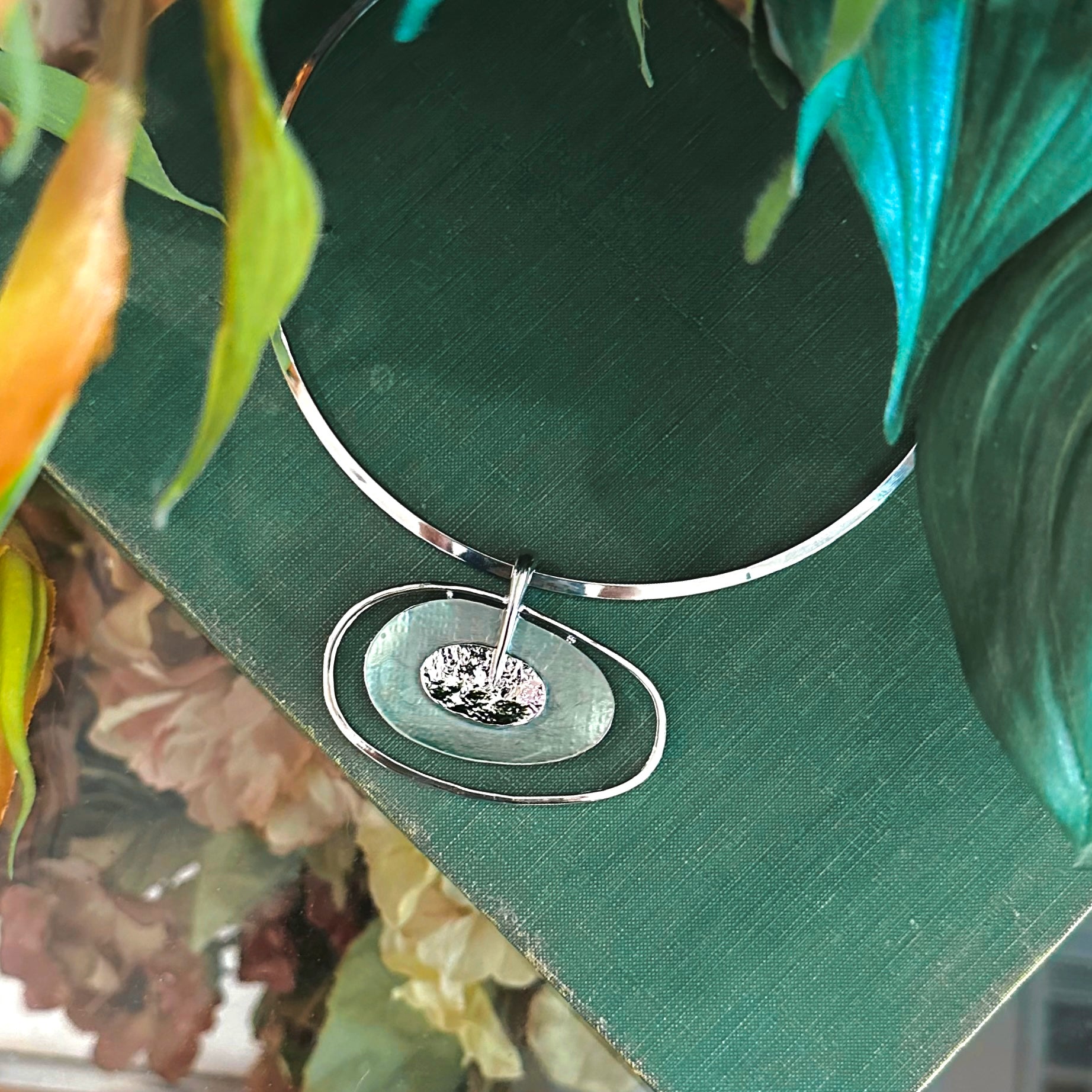 rhodium plated round choker with an Open Oval Pendant with two textured overlapping oval disks in the center with the bottom disk painted in green enamel rests on a green book with leaves in the foreground