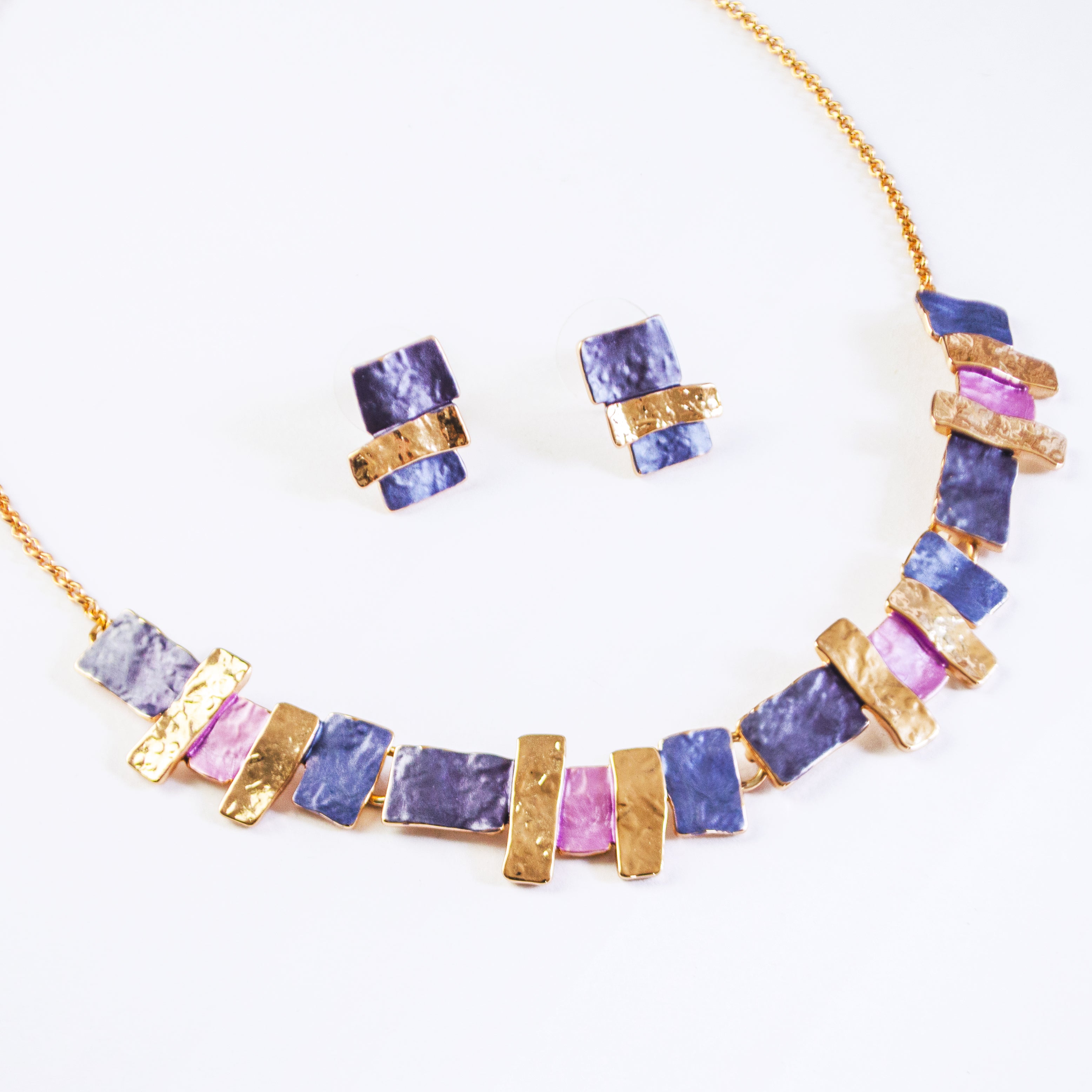 Pair of rose gold plated earrings with three textured stacked squares and matching necklace with rose gold, purple and pink square and rectangle pendants against a white background