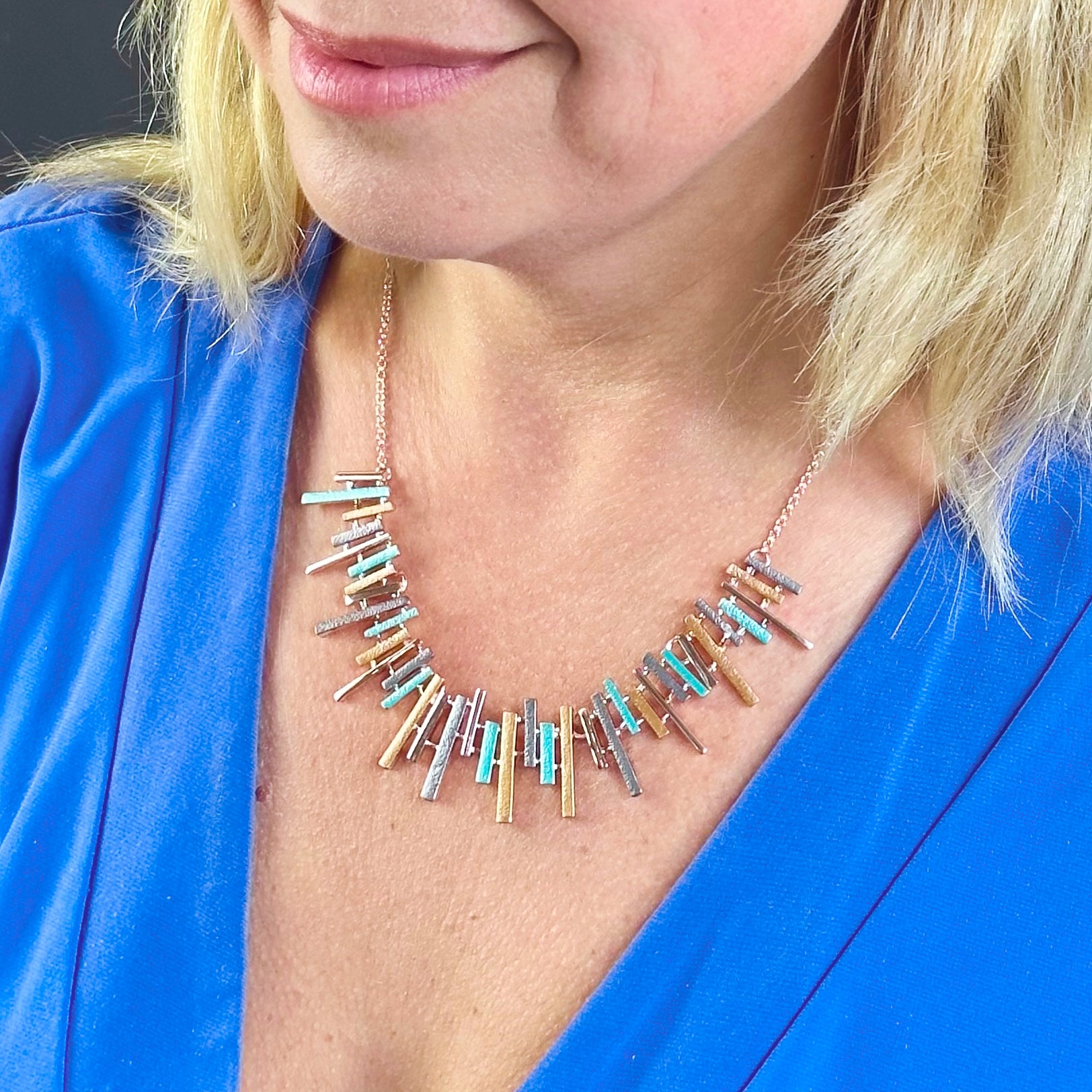 Adjustable Rose Gold Plated Necklace with a cable chain and connected pendants with vertical bars of varying lengths painted in teal, gray, and gold enamel on model wearing blue blouse