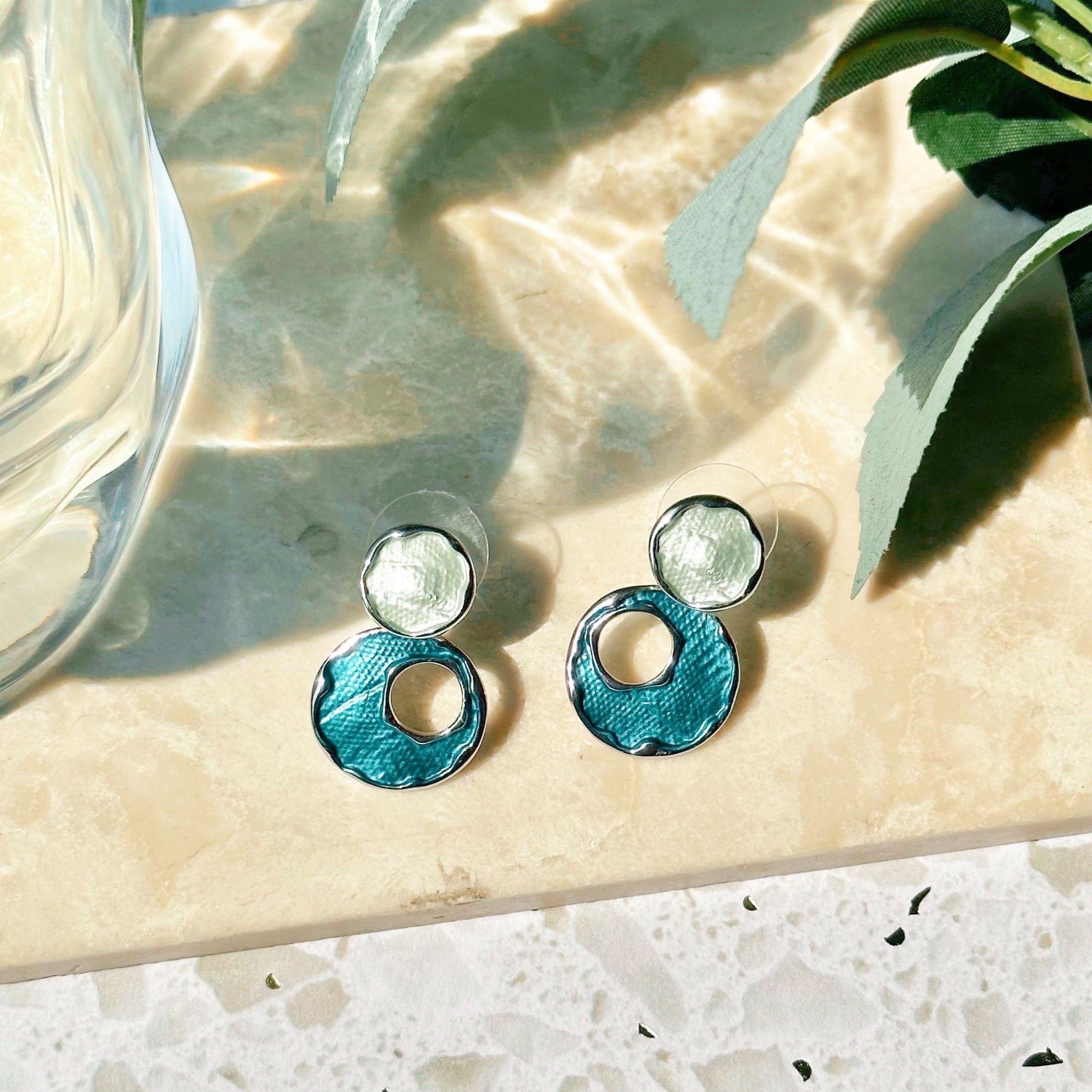 A pair of rhodium plated stud earrings made with two circles, the top painted with silver enamel, and the bottom larger circle painted in blue enamel with an off-center circle cutout rest on a marble tile with leafy plants on a white stone background