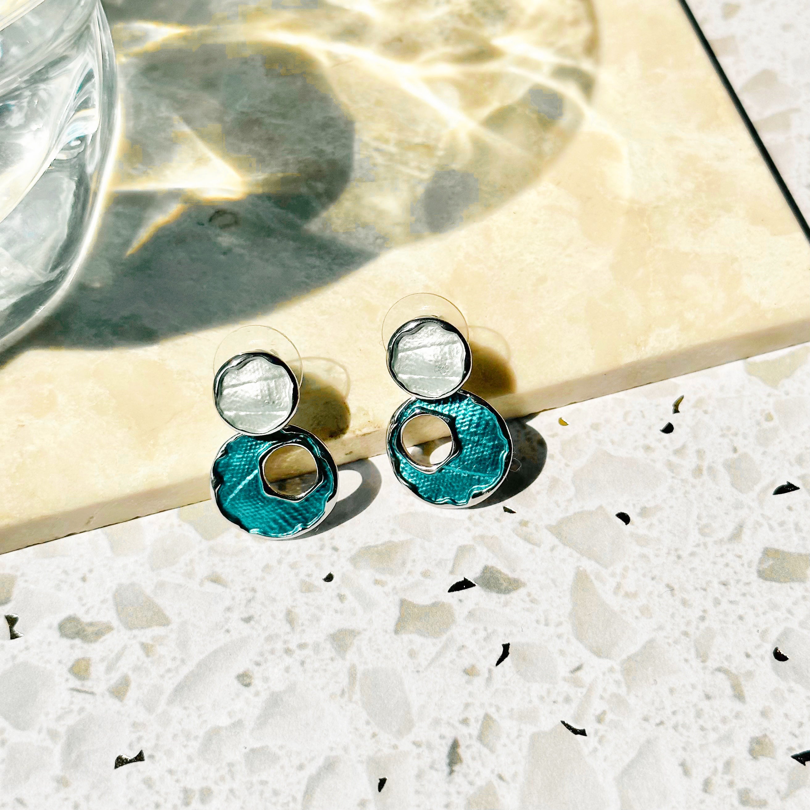 A pair of rhodium plated stud earrings made with two circles, the top painted with silver enamel, and the bottom larger circle painted in blue enamel with an off-center circle cutout rest on a marble tile with leafy plants on a white stone background