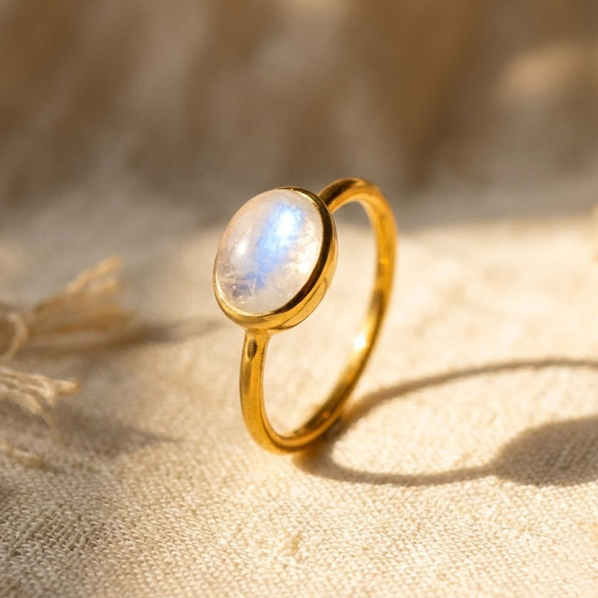 Oval rainbow moonstone ring in yellow gold vermeil with a glowing blue flash on a warm neutral background.