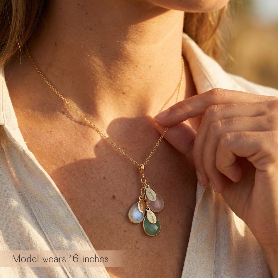 A person in a beige shirt wears a cascading necklace with birthstones and name tags visible. The necklace features the October birthstone, the July birthstone and the April birthstone. 