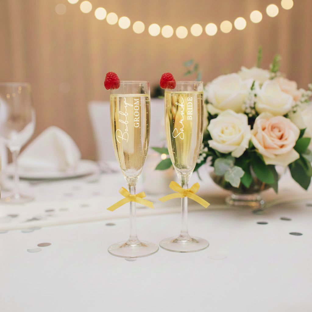 Personalized bride and groom champagne flutes with names engraved, topped with raspberries and tied with gold bows, displayed on an elegant wedding reception table with roses and soft string lights; custom engraved wedding toasting glasses gift for couple.