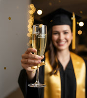 Smiling graduate in cap and gown holding personalized champagne flute, custom engraved with name and class year, gold tassel and bokeh lights in background, celebratory graduation champagne glass gift for high school or college senior party and keepsake.