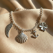 Silver anklet with various charms including a shell, anchor, and clover on a beige fabric background. The dainty sterling silver anklet features carefully spaced miniature charms along a slender chain, offering a refined yet playful aesthetic that makes it a perfect personalized gift for bridesmaids, sisters, or best friends.